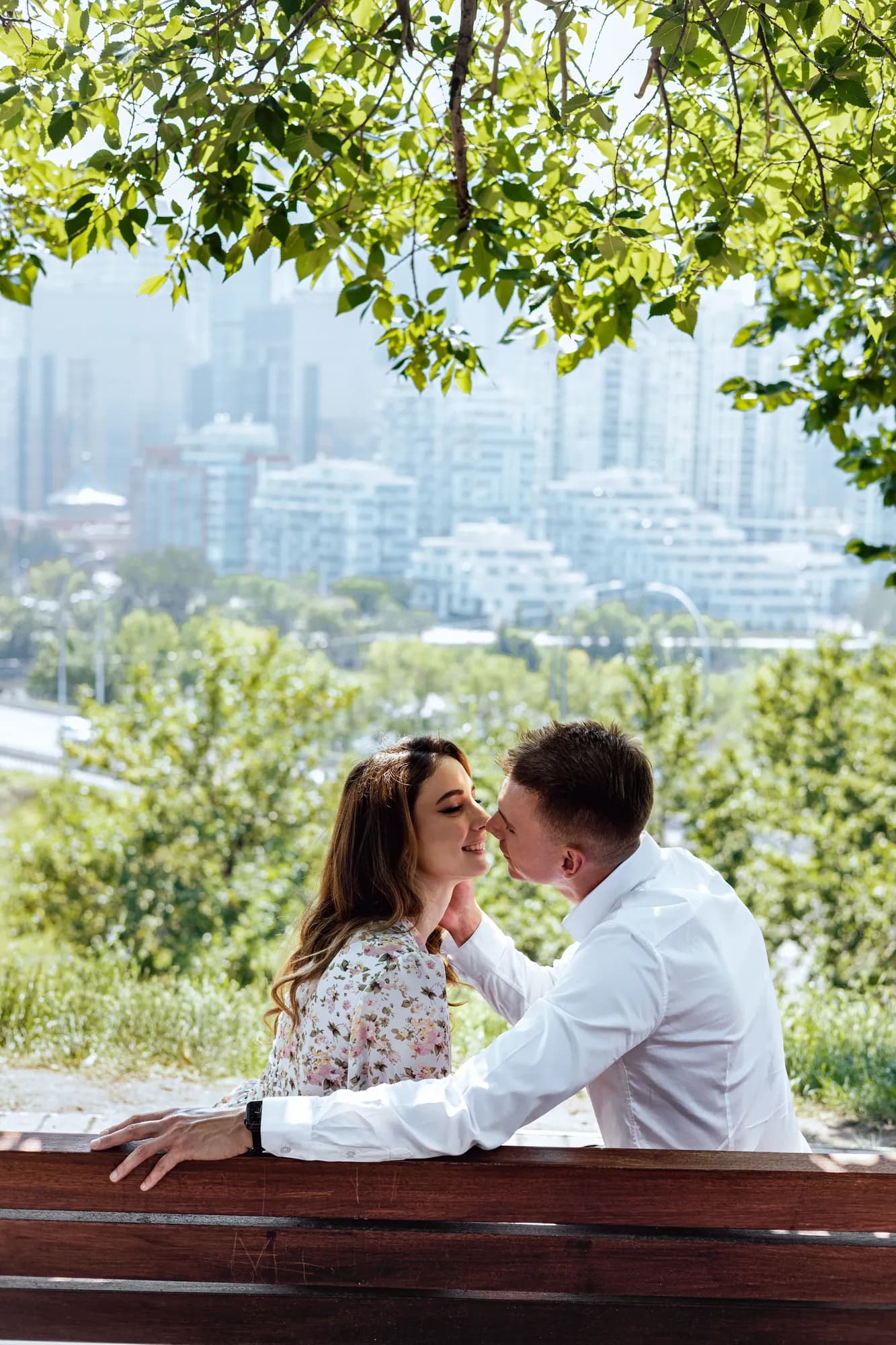 A couple sitting close on a bench with the city skyline behind them
