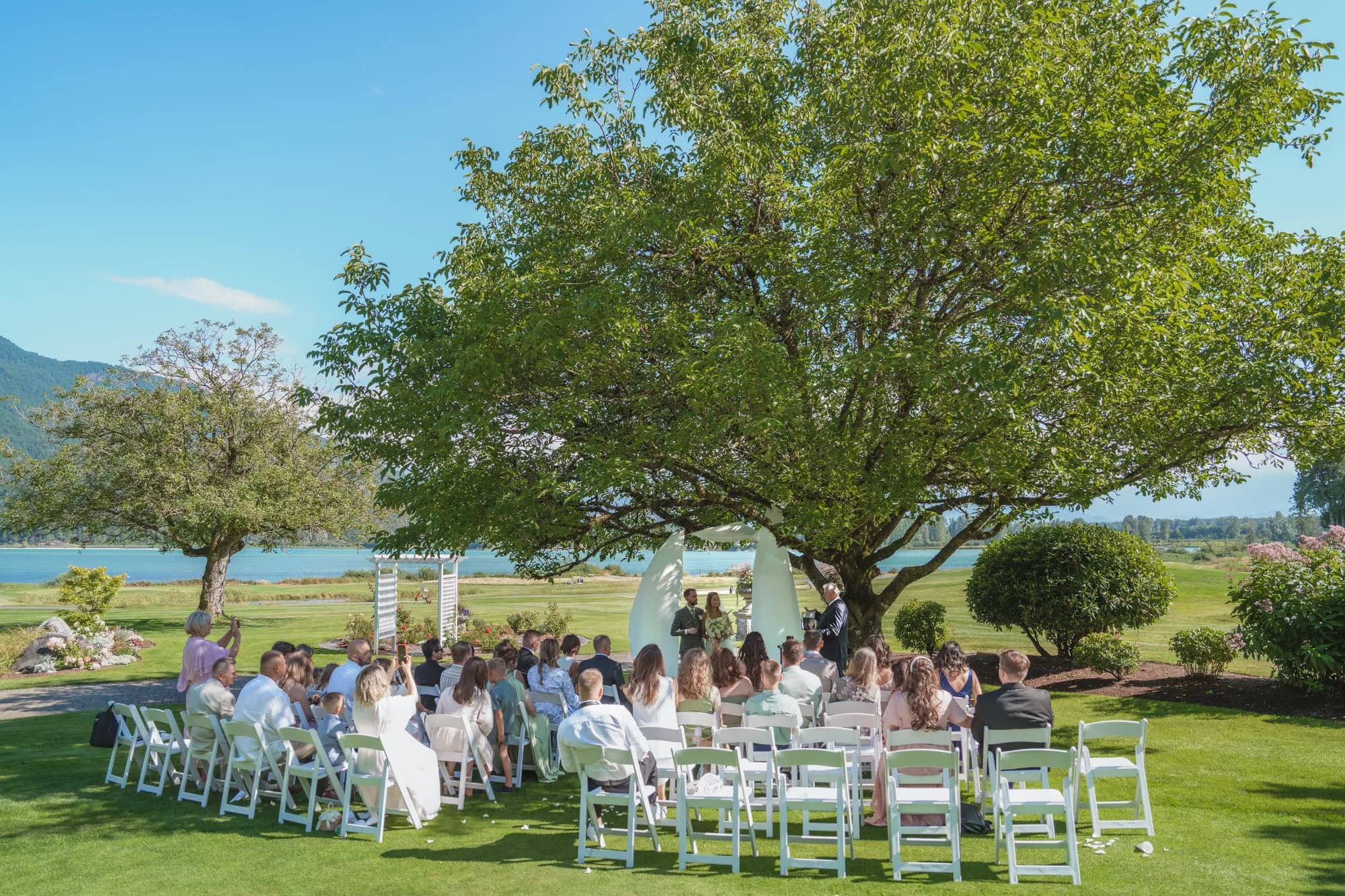 An outdoor ceremony beneath a large tree, guests seated facing the lake