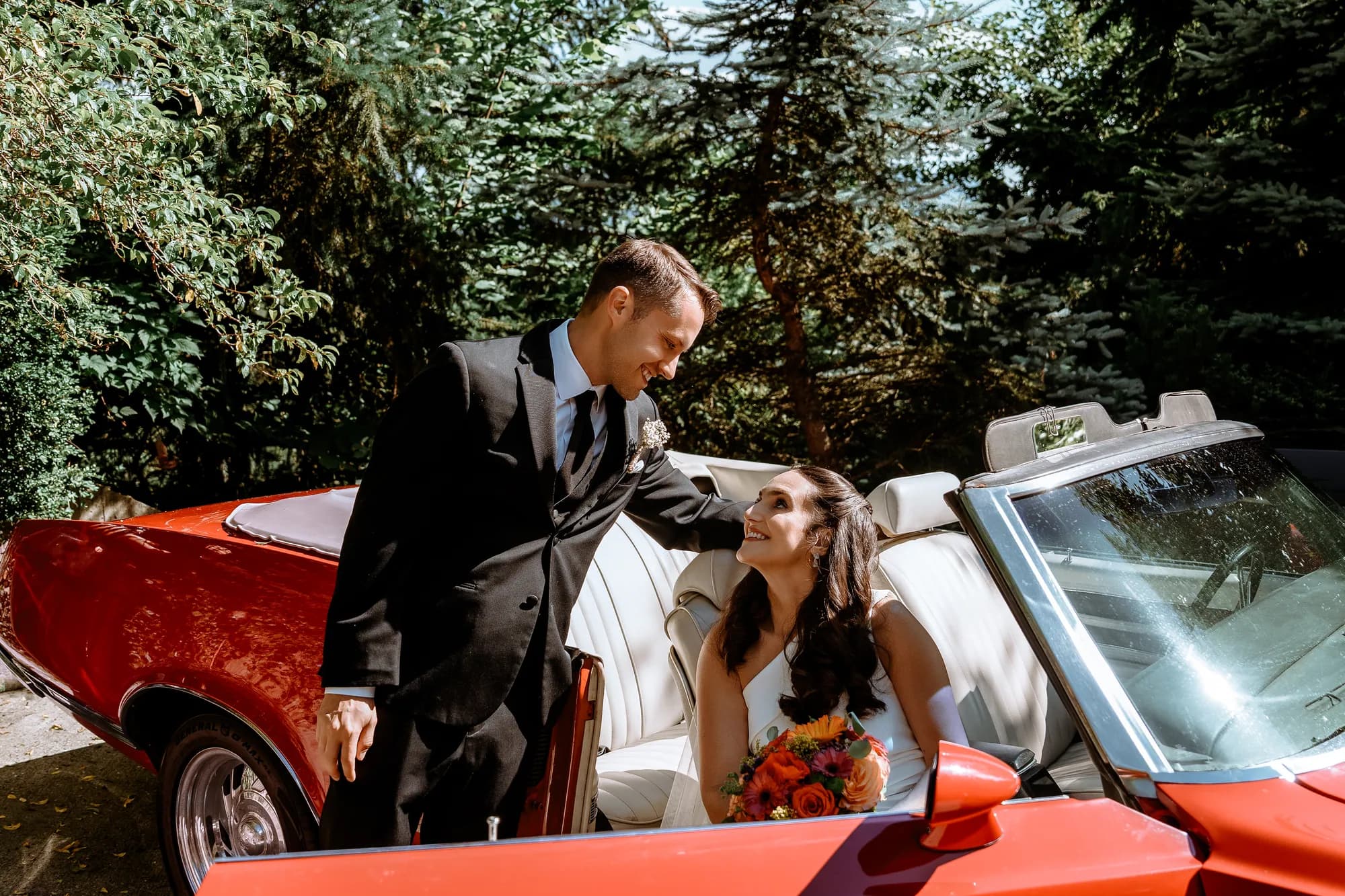 A groom leaning over the door of a red vintage convertible, smiling down at his bride in the passenger seat holding a bright bouquet