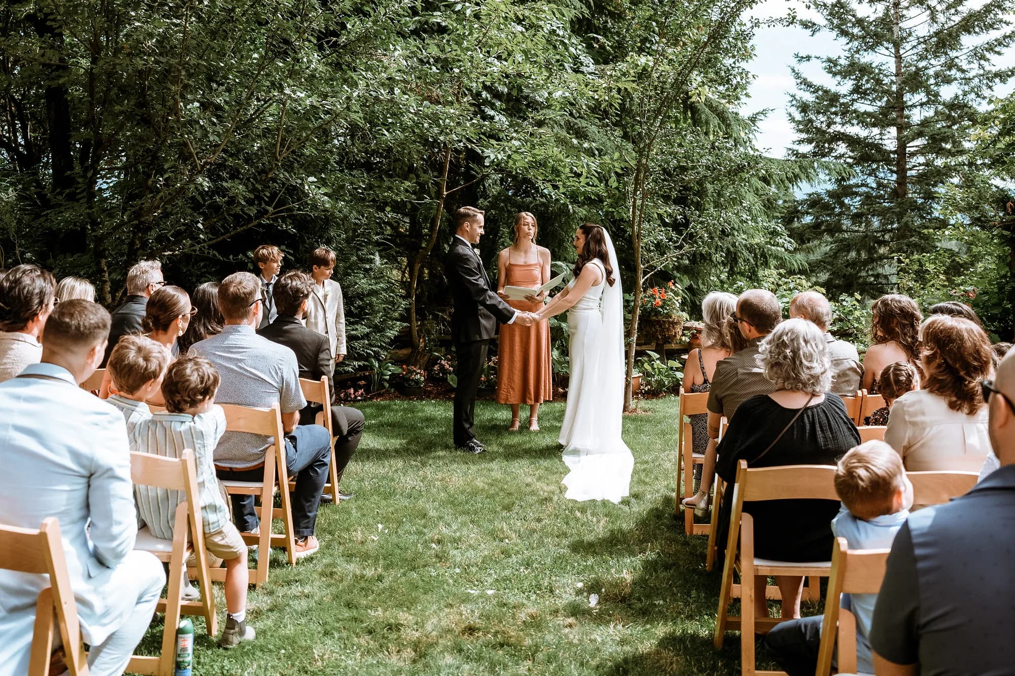 A forest ceremony in progress, guests seated on wooden chairs facing the couple